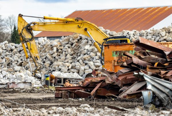 Excavator with hydraulic breaker beside a steel skip and piles of twisted girders and concrete rubble on a demolition jobsite